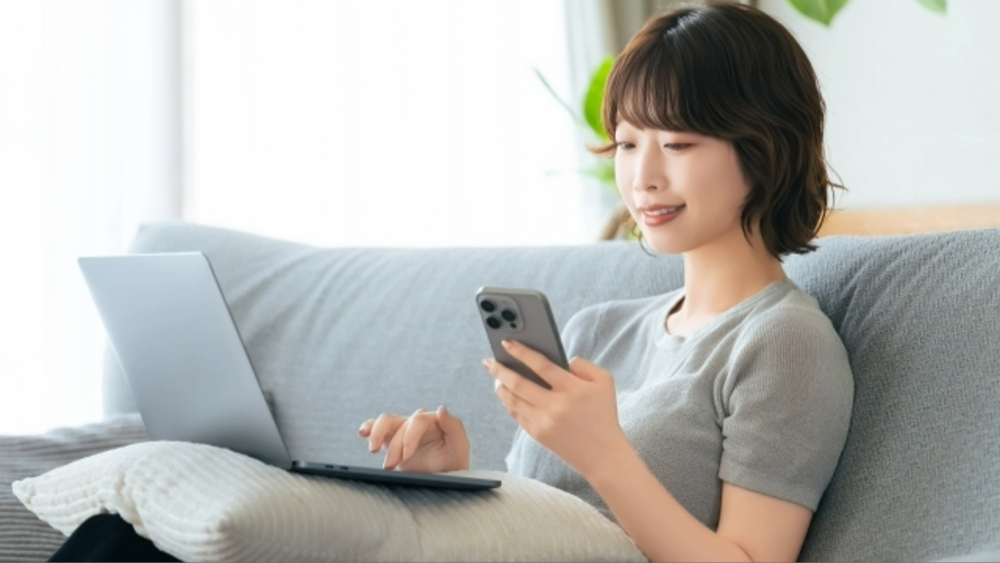 オンラインでの動画接客やチャットサポートを利用しながら、自宅のソファでリラックスしてスマートフォンとパソコンを併用する女性の様子。