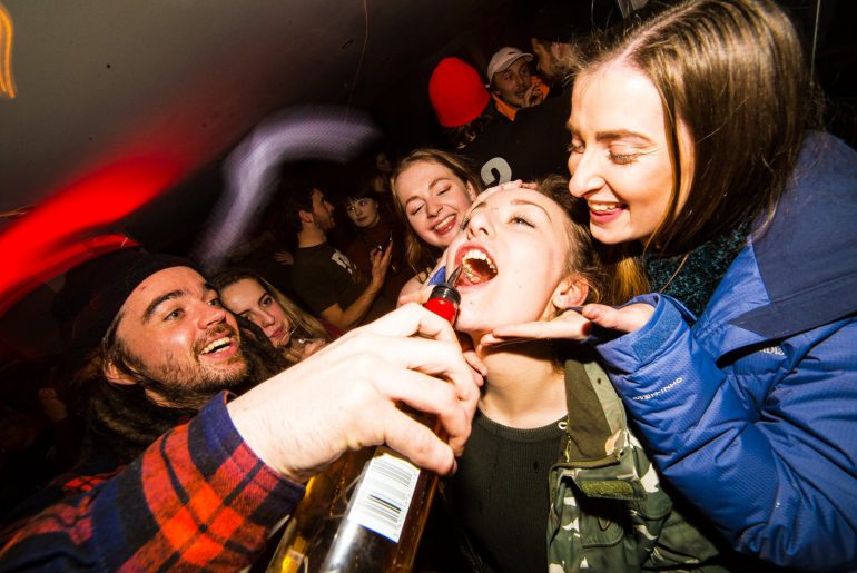 man pouring bottle of alcohol into woman's mouth at apres ski party