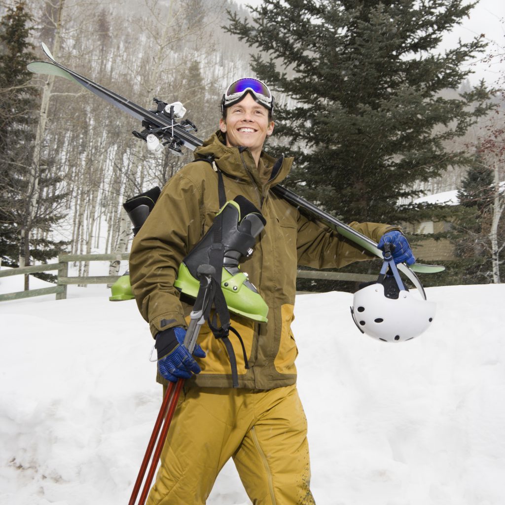 Attractive man in winter clothing walking in snow carrying ski equipment and smiling.