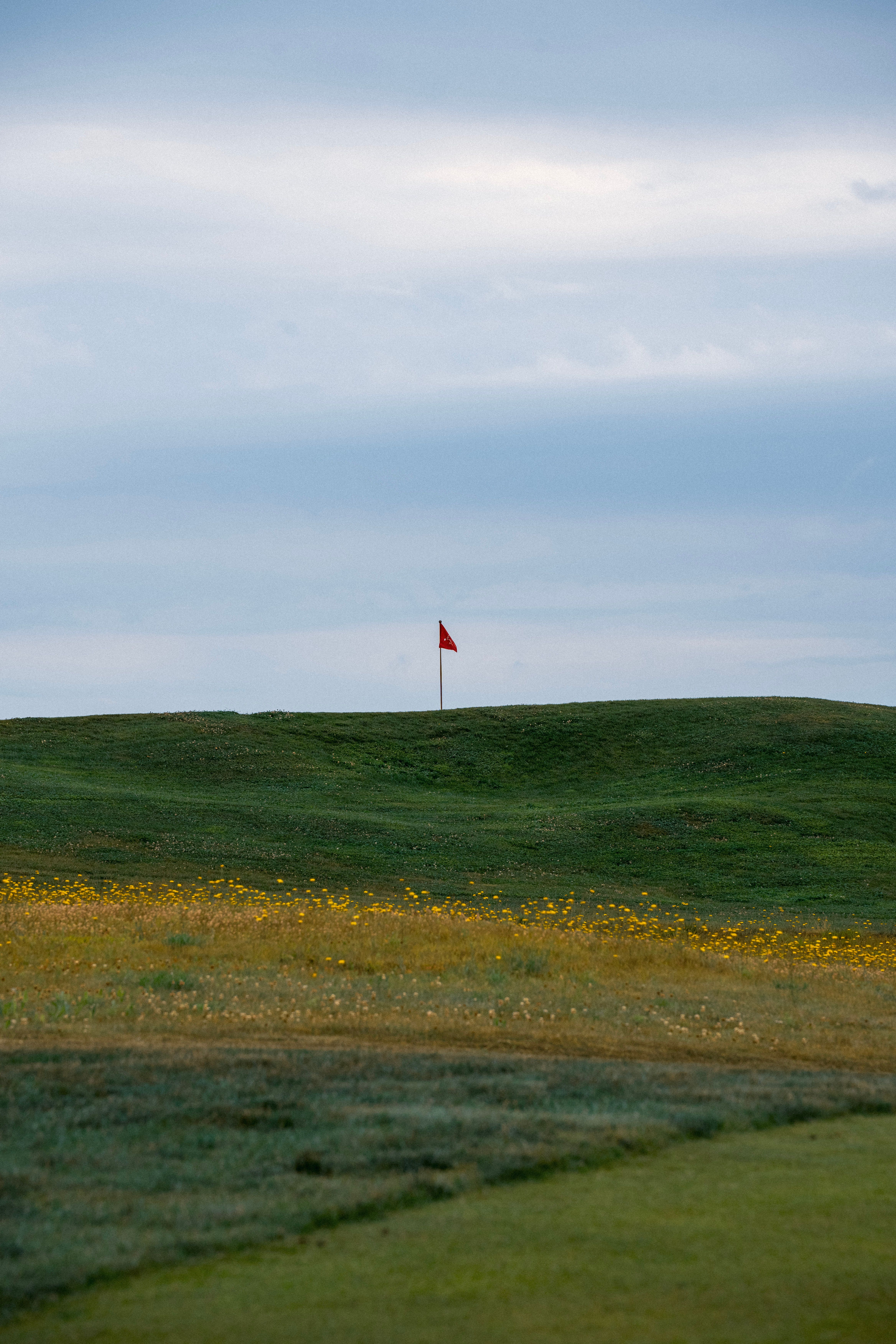 A red golf flag stands atop a vibrant green hill under a cloudy sky.