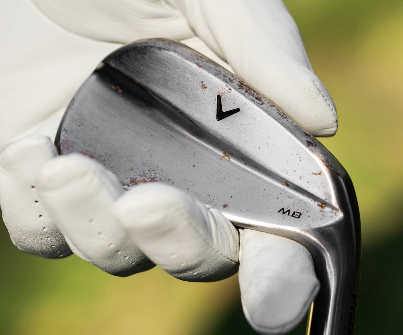 A gloved hand holds a silver golf club head with a distinct black 'V' logo.