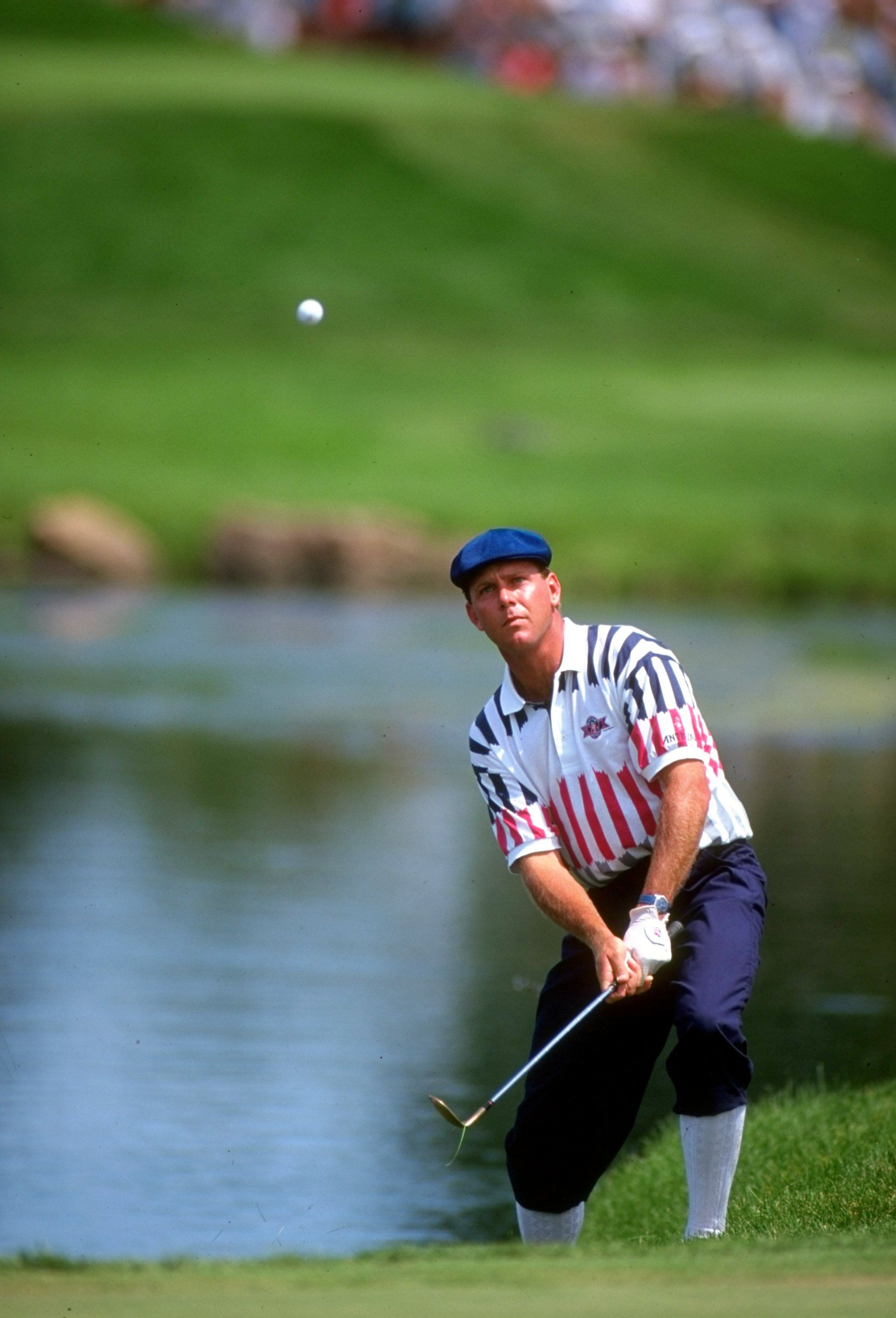Golfer in a patriotic shirt hits a golf ball over water on a green course.