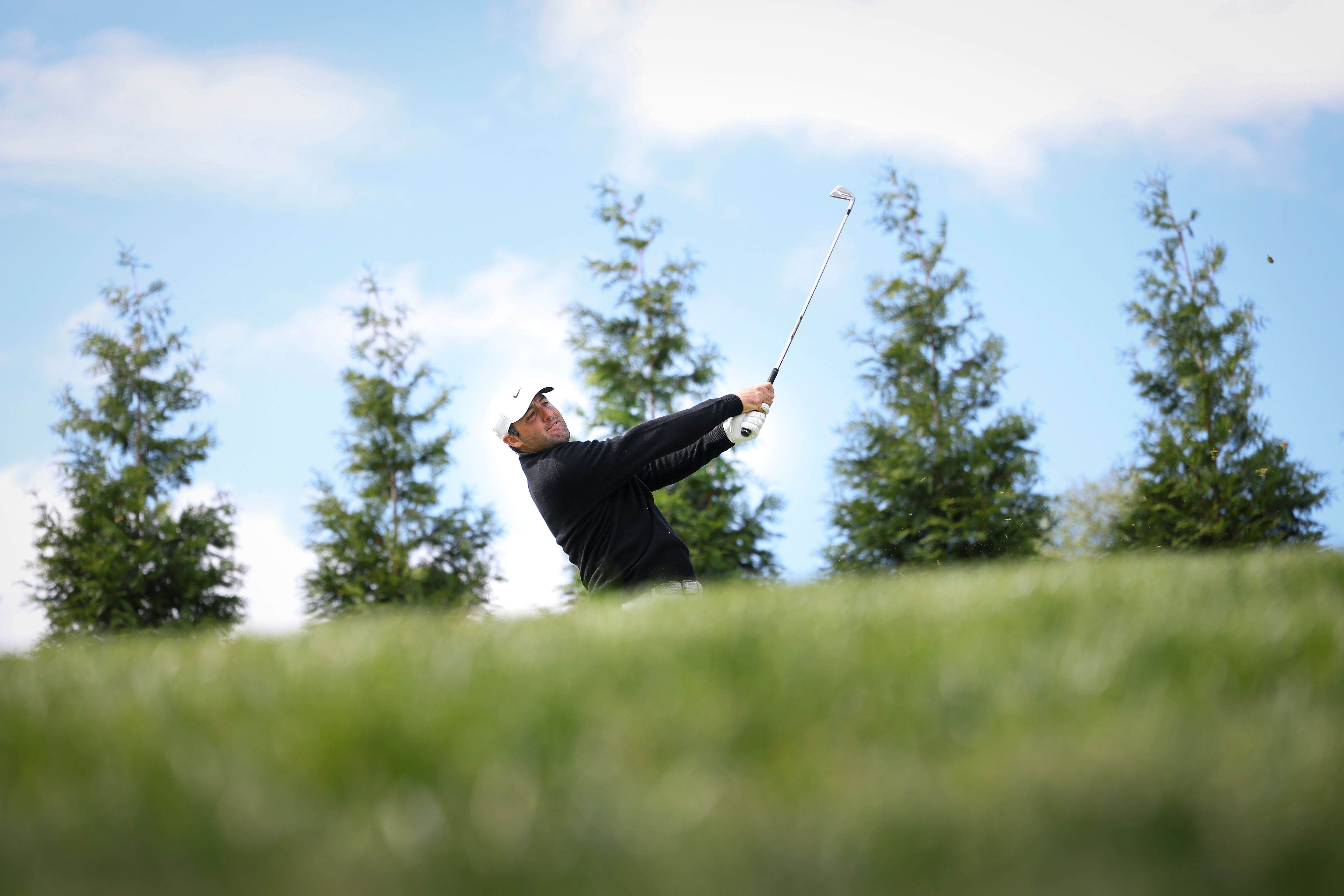 Golfer swings club on green hill under a blue sky with several trees.