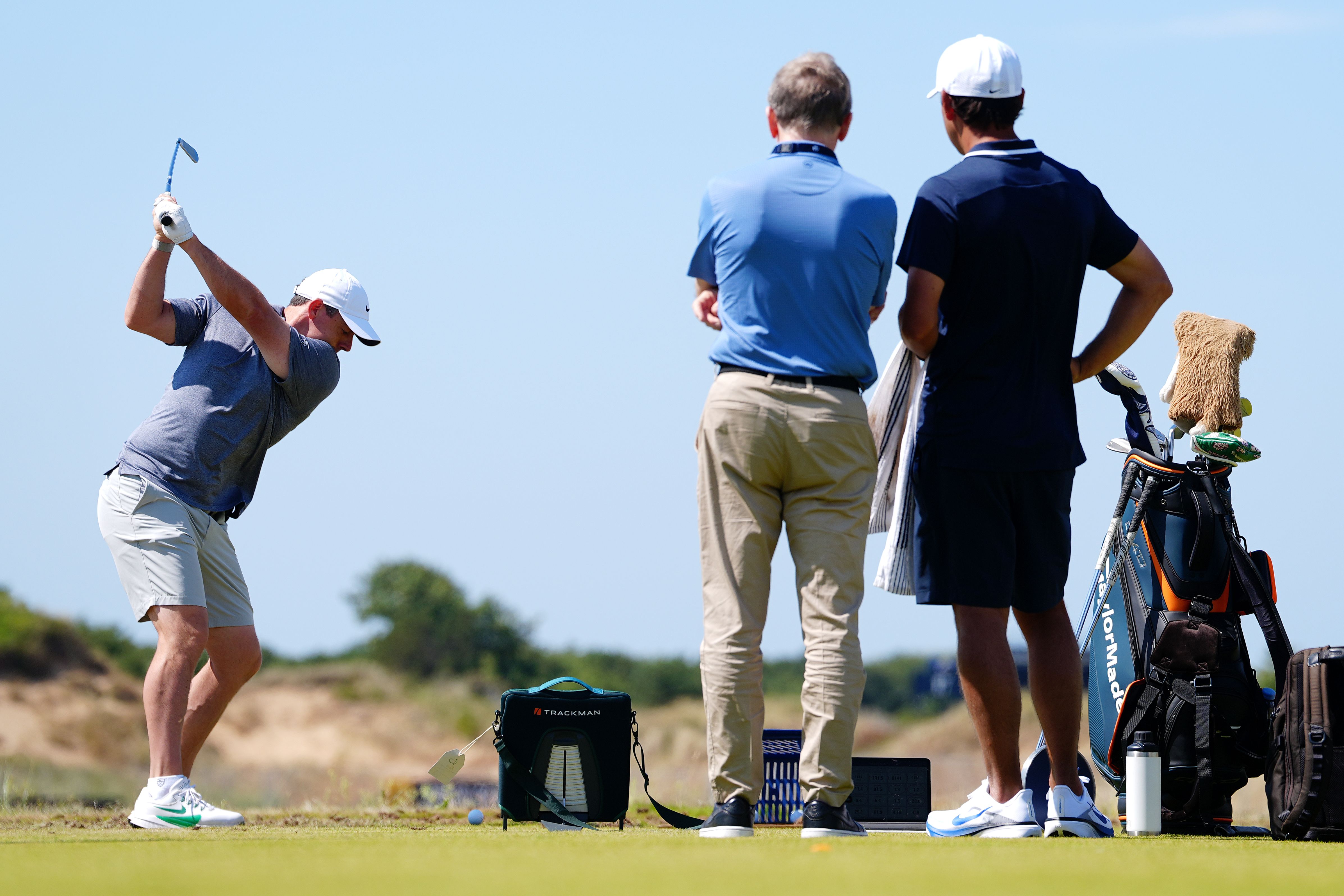 Golfer in grey shirt swings club as two men watch on a sunny course.