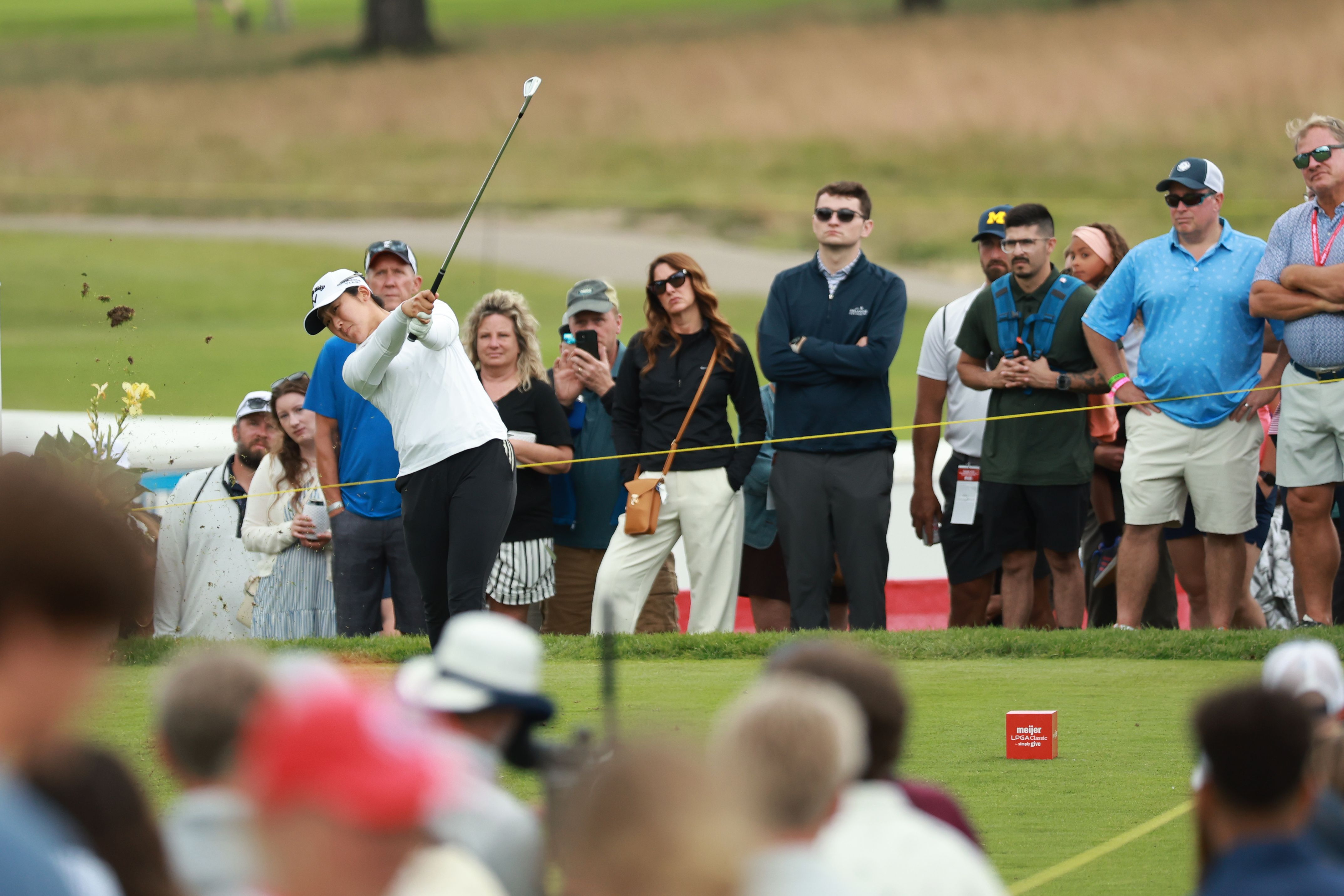 Golfer mid-swing on a green course, the ball flying, watched by a blurred crowd.