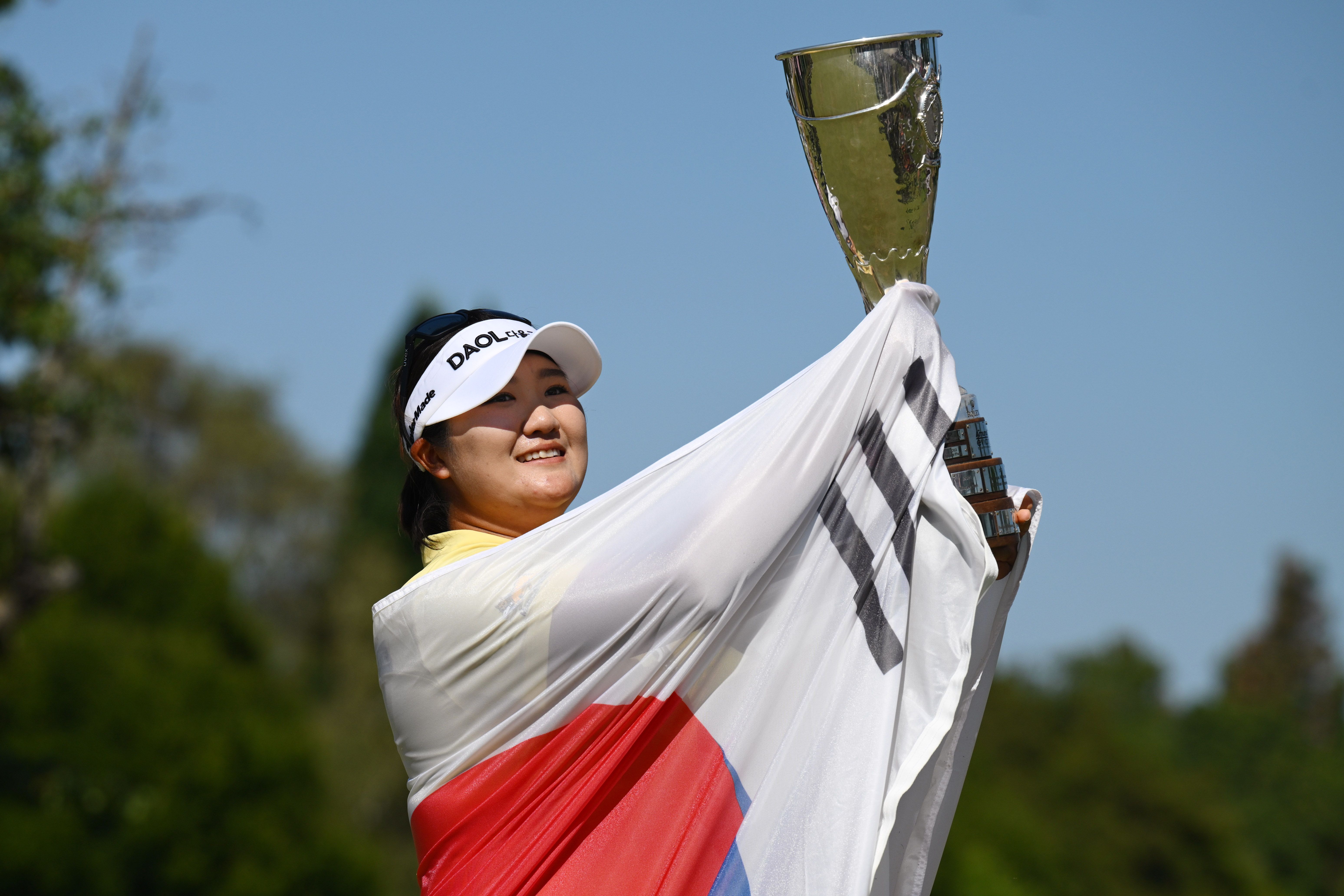 A happy female golfer holds a gold trophy and a South Korean flag overhead outdoors.