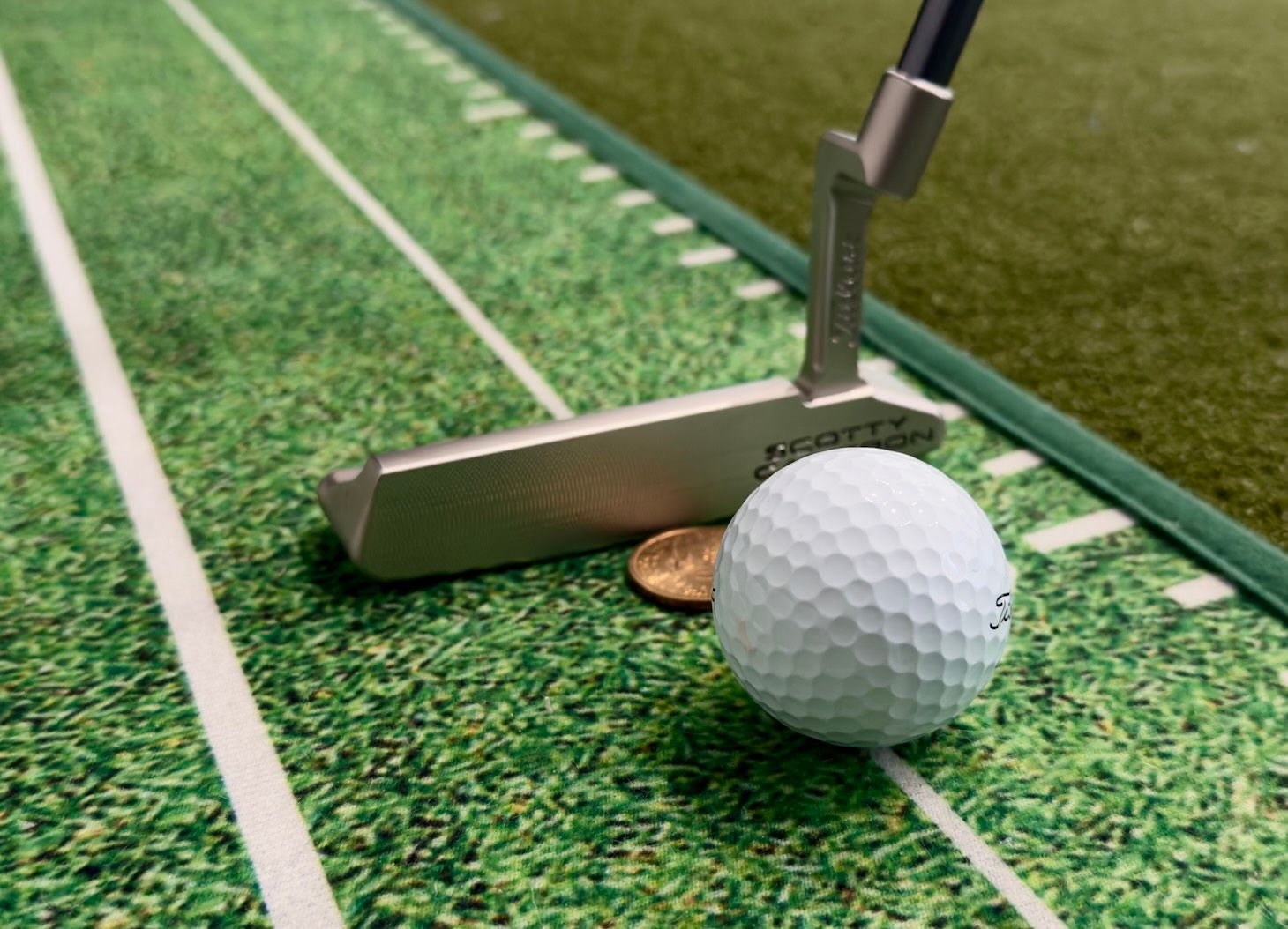 A golf ball and putter rest on a green putting mat with a coin marker.