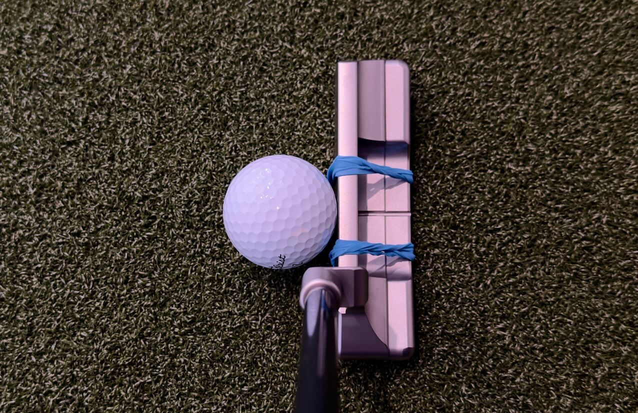 A golf putter with blue elastic bands and a white golf ball on a green mat.