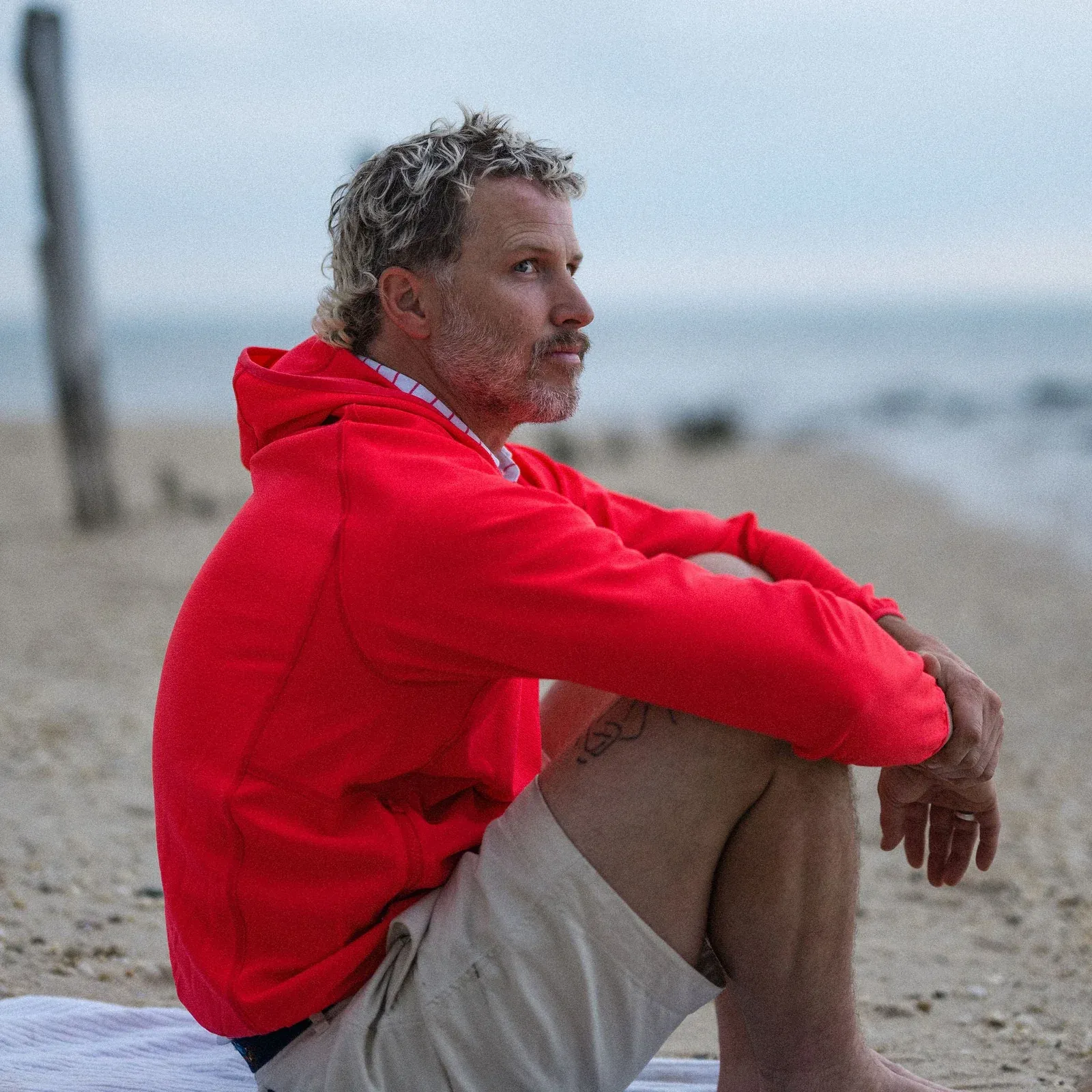 Man in a red hoodie sits contemplatively on a sandy beach looking at the ocean.