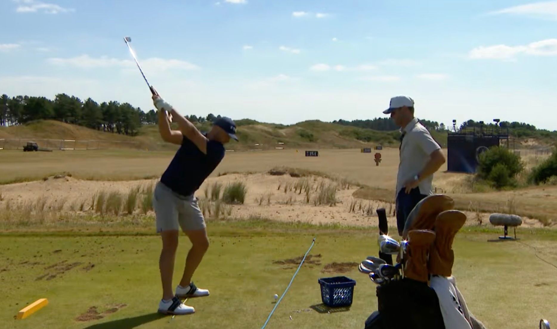Golfer in mid-swing on a sunny course with an instructor watching closely.