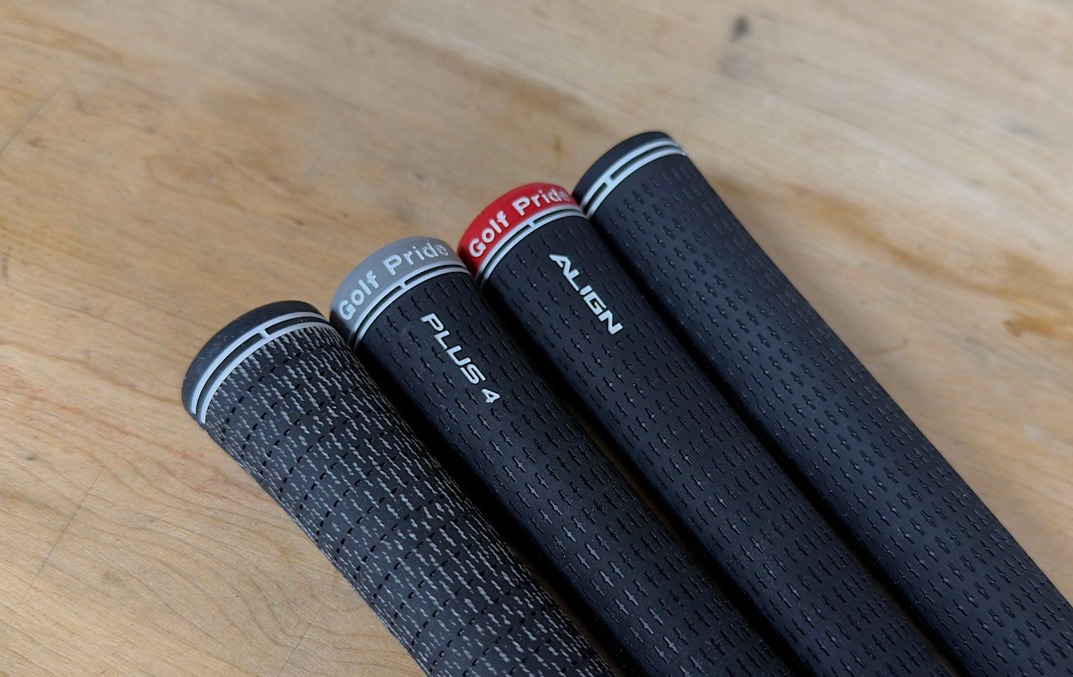 Four different black golf club grips lie diagonally on a light wooden surface.