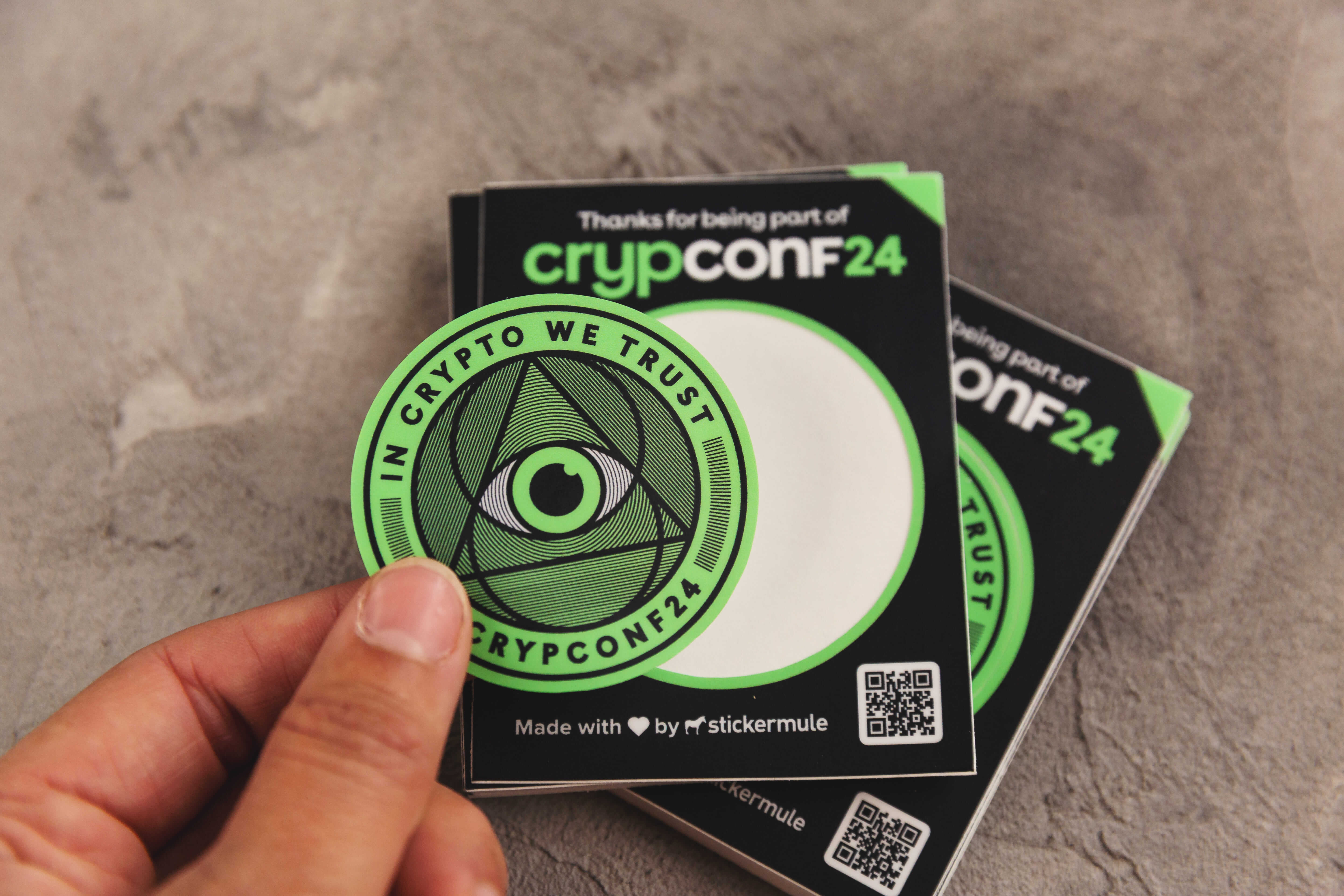 Sticker semi-troquelado verde brillante de una convención empresarial sobre criptografía, con una mano sujetando el sticker despegado