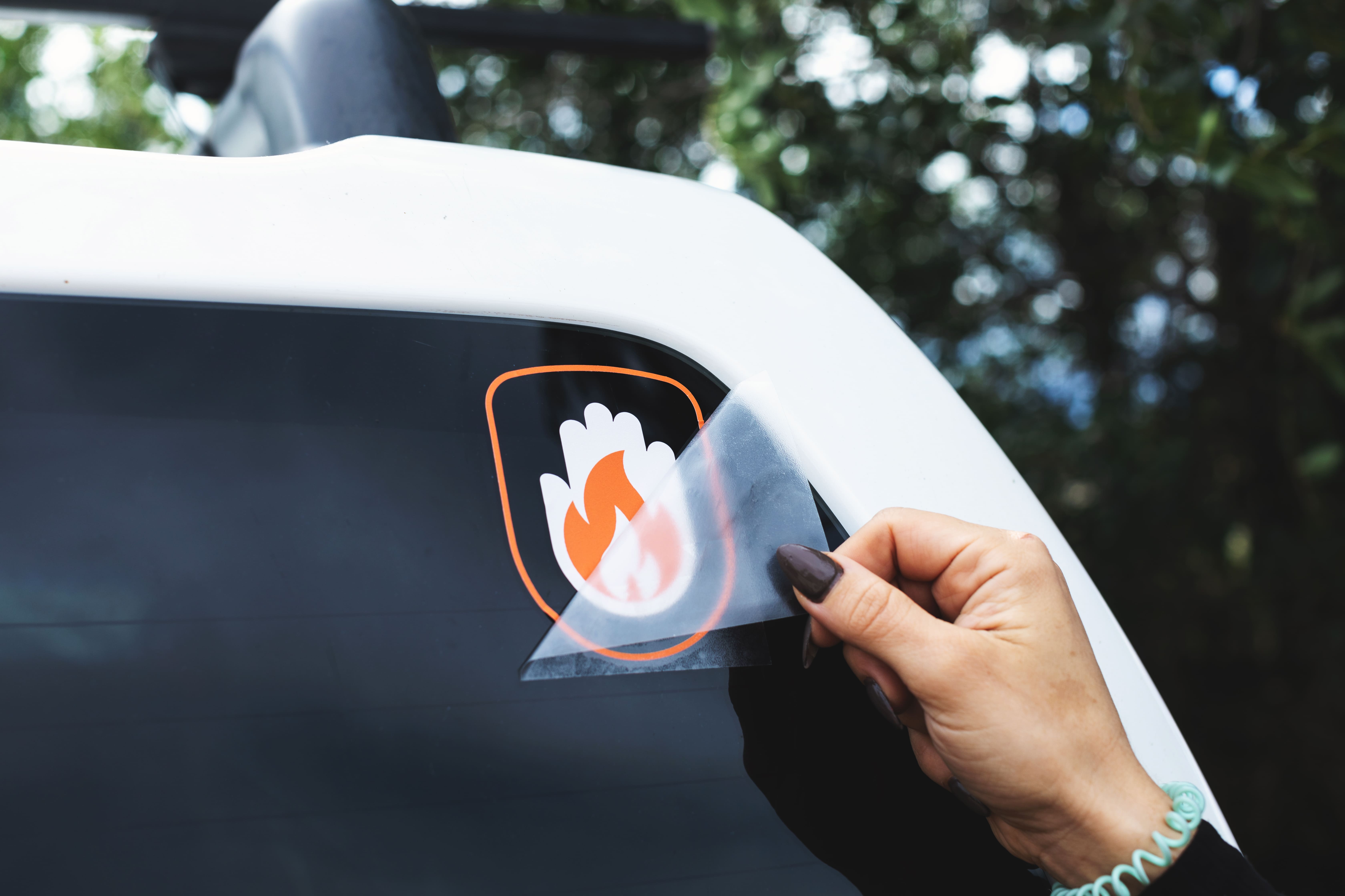 Sticker transferible a color en blanco y naranja de una mano combinada con una llama, aplicada a la luna trasera de un coche blanco. Una mano despega la hoja de transferencia del sticker