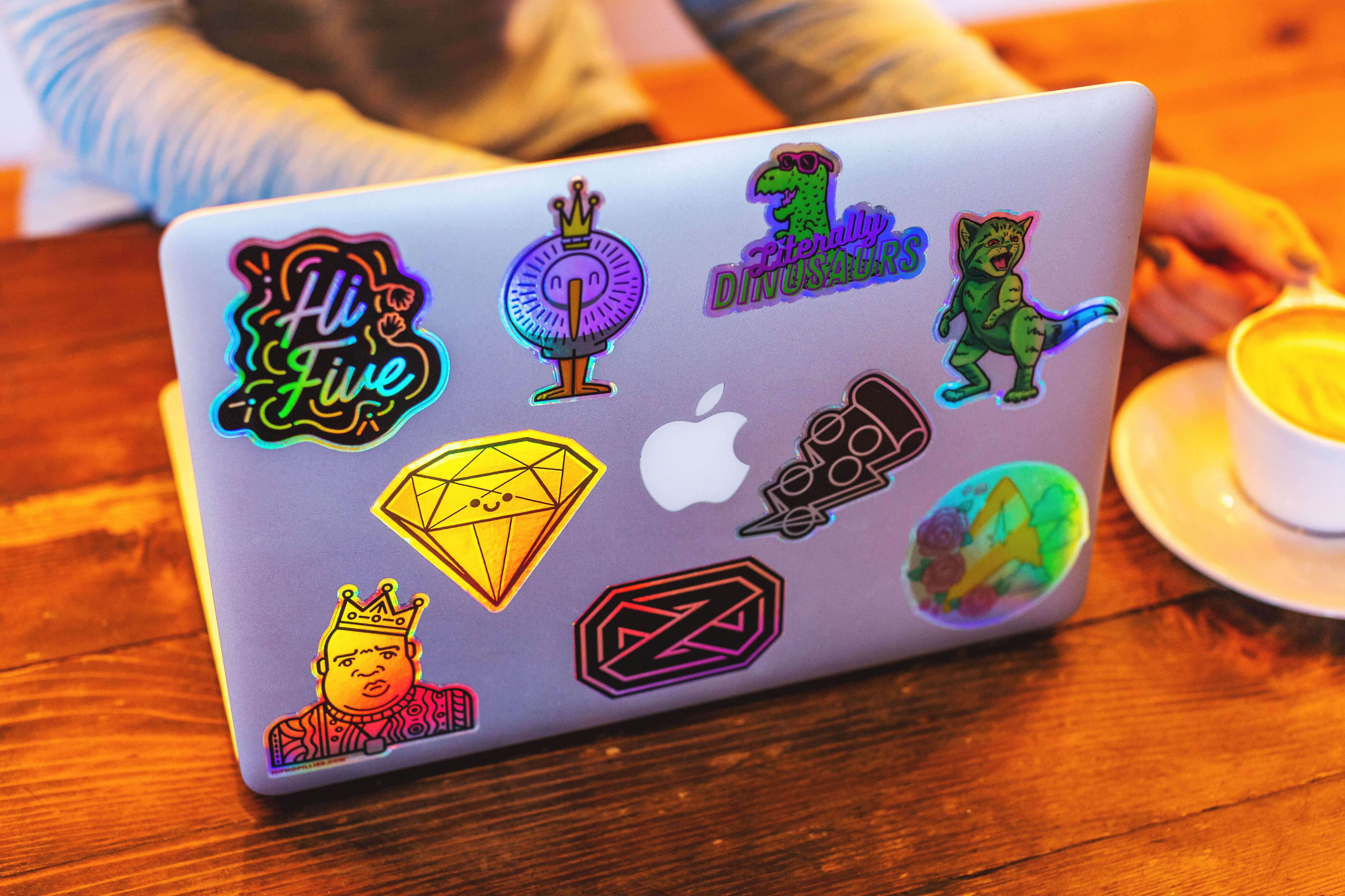 Laptop decorada con stickers troquelados coloridos, incluyendo diseños de dinosaurios, coronas, gemas y un sticker holográfico de High Five.
