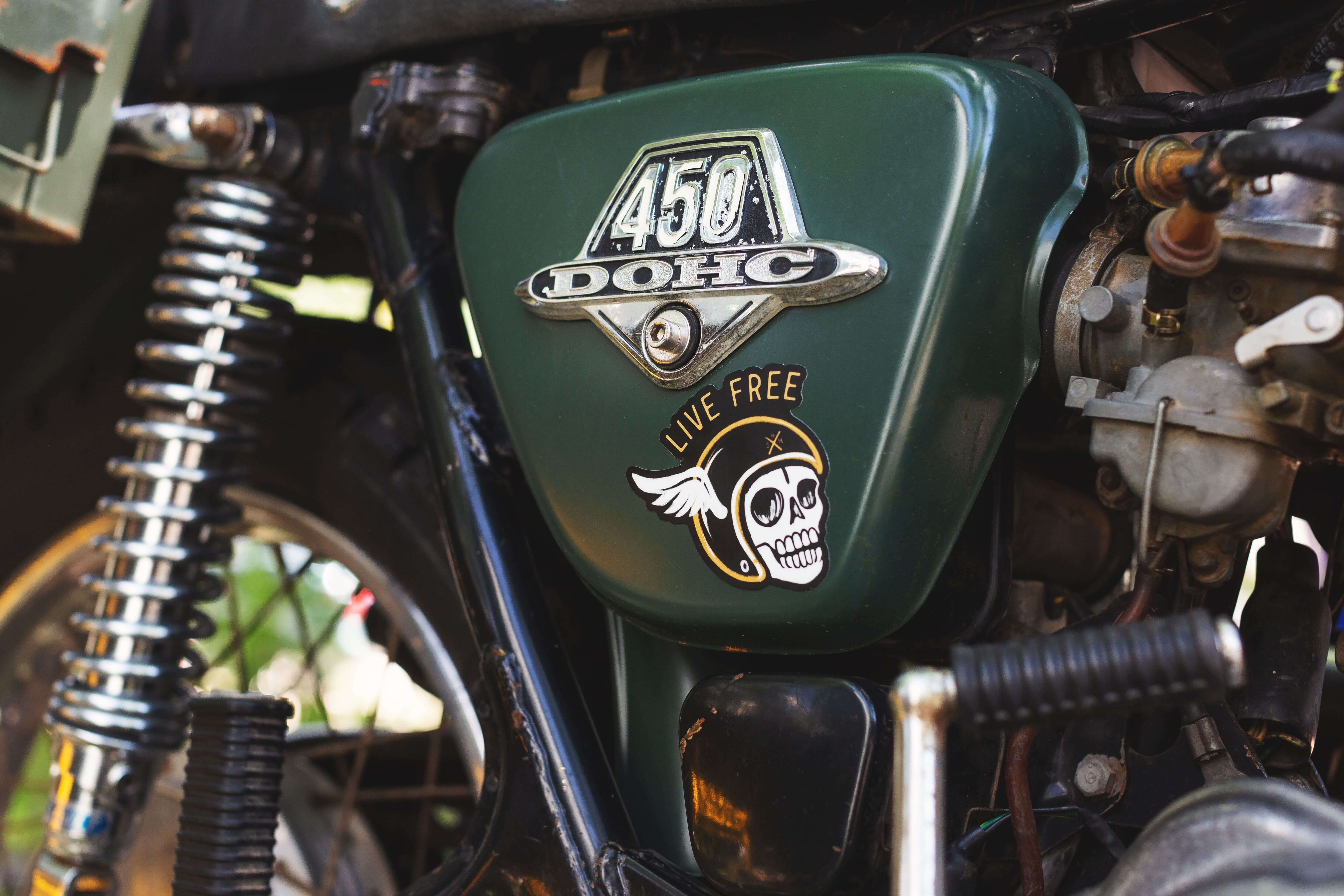 Sticker troquelado 'Live Free' en un estilo de motocicleta, mostrando un cráneo con un casco de motor, pegado en el tanque de una motocicleta vintage verde.