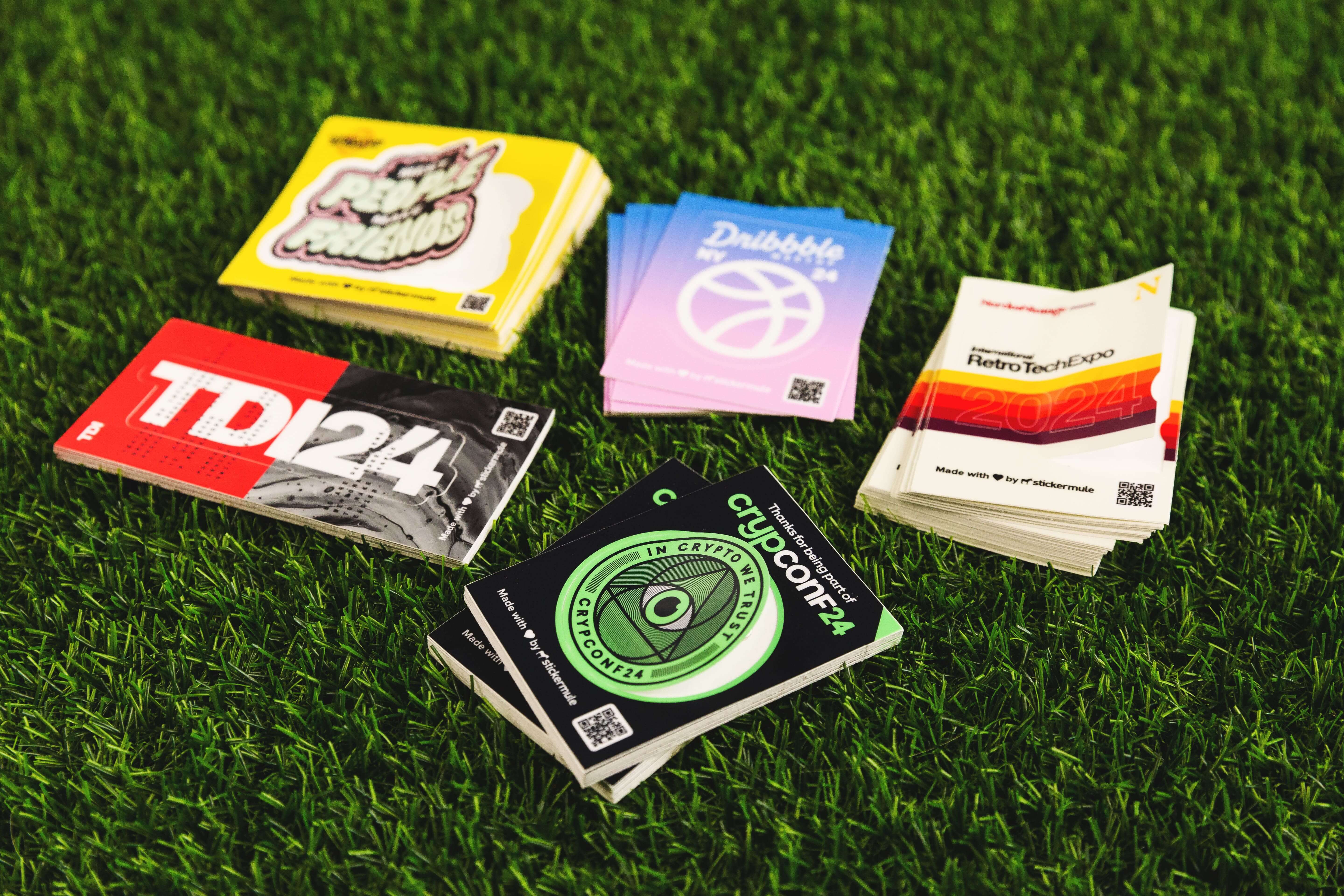 Pilas de stickers únicos de eventos sobre césped, con diseños audaces y personalizados para marcas como CryptoConf, Dribbble y RetroTechExpo.