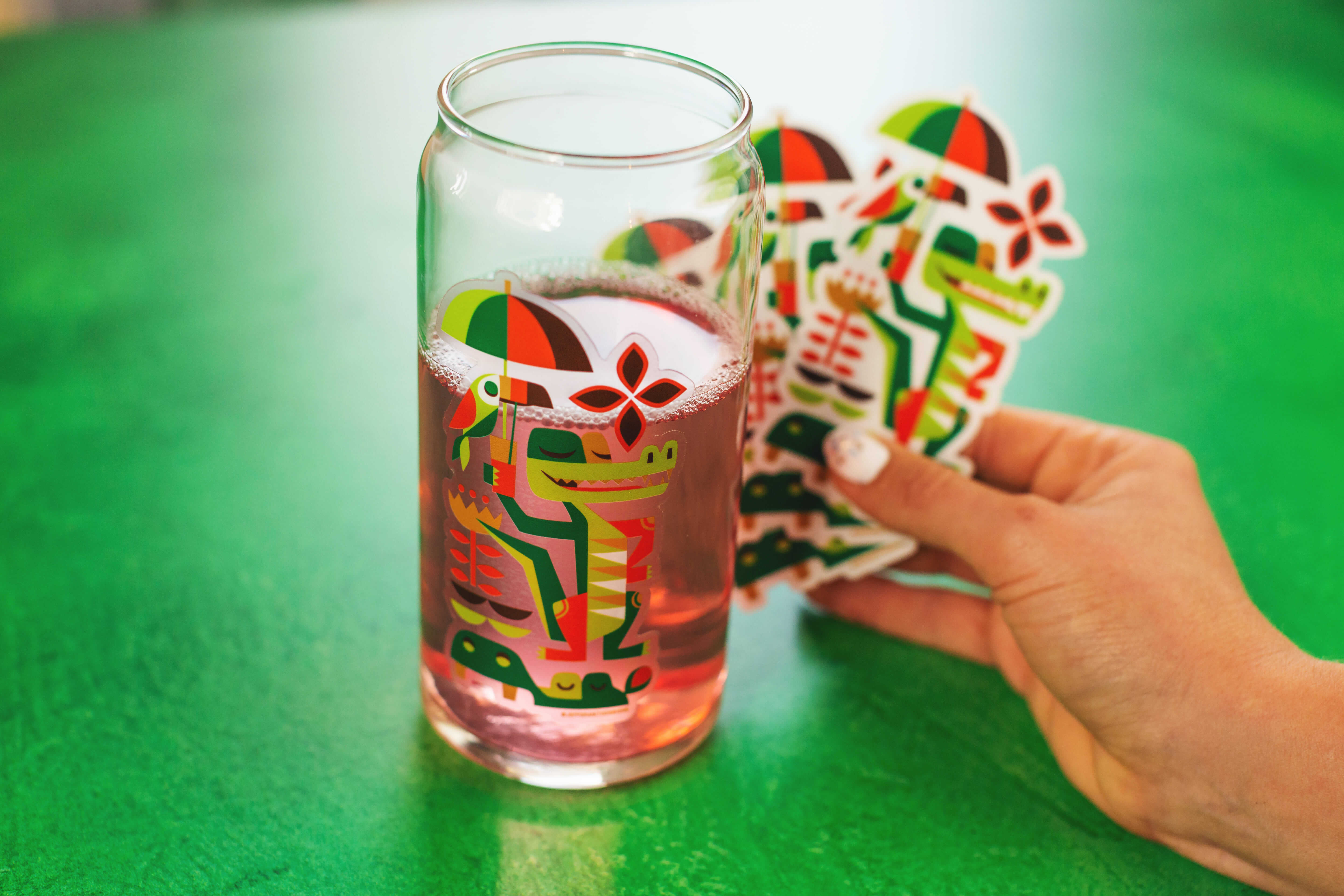 A clear sticker of a vector illustration of a crocodile holding a sun umbrella applied to a glass of lemonade.