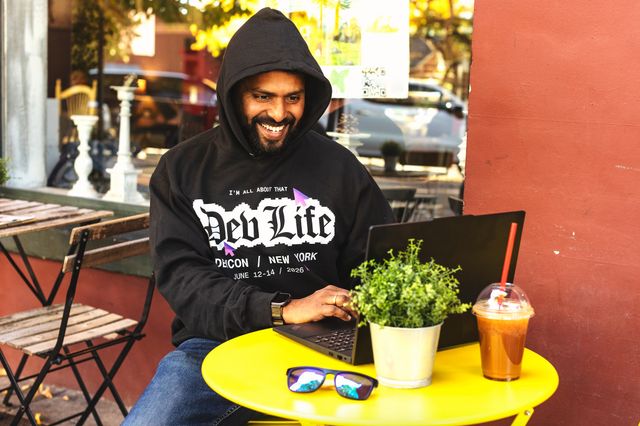 man in a black hoodie with a graphic on the front typing on a laptop at an outside cafe