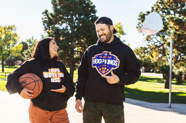 a man and a woman talking on a basic ball court each wearing a black hoodie with a graphic on the front. 