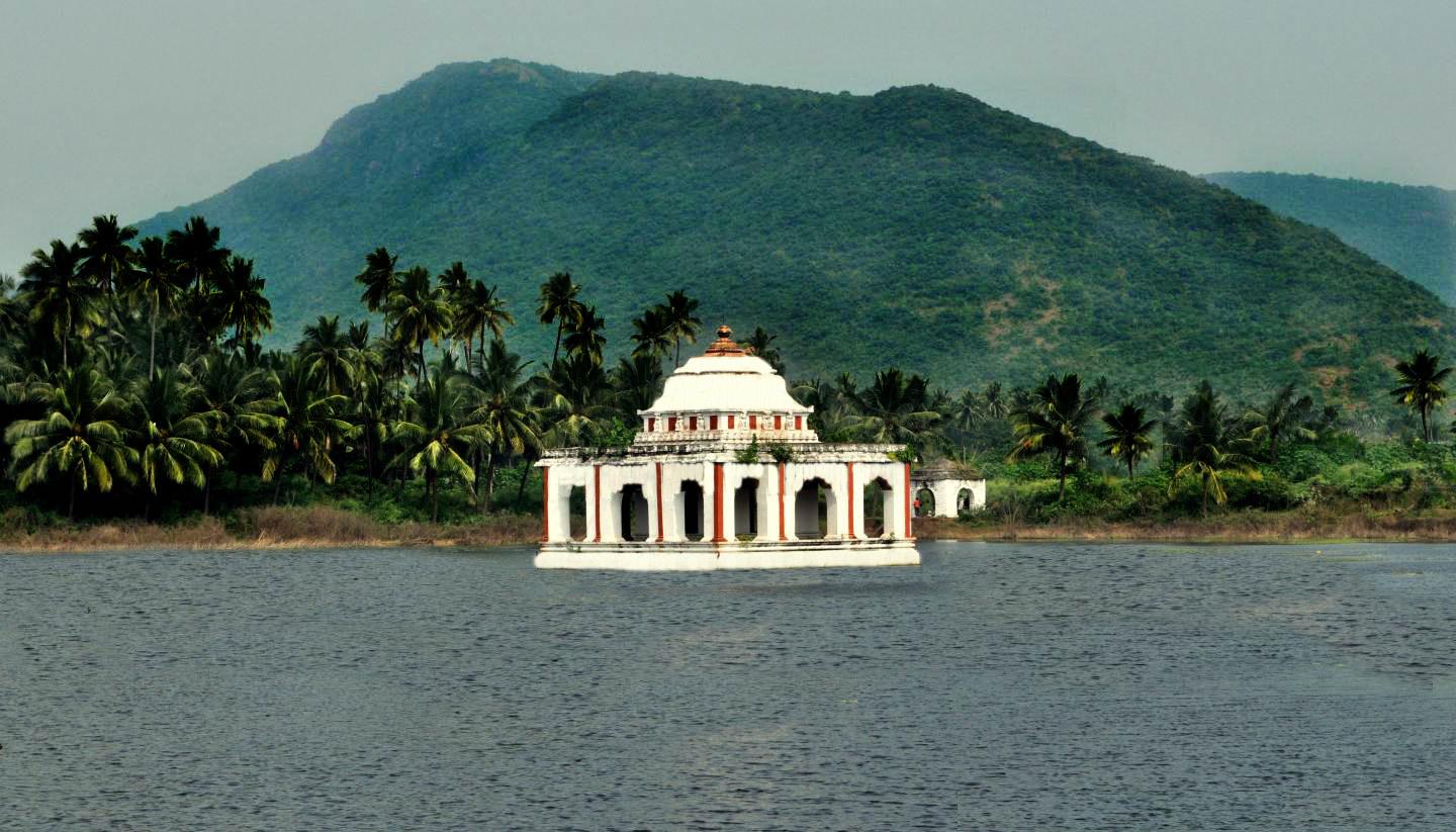 A view of a pond with a hall like structure built in the middle, surrounded by green trees and the hills in the background.