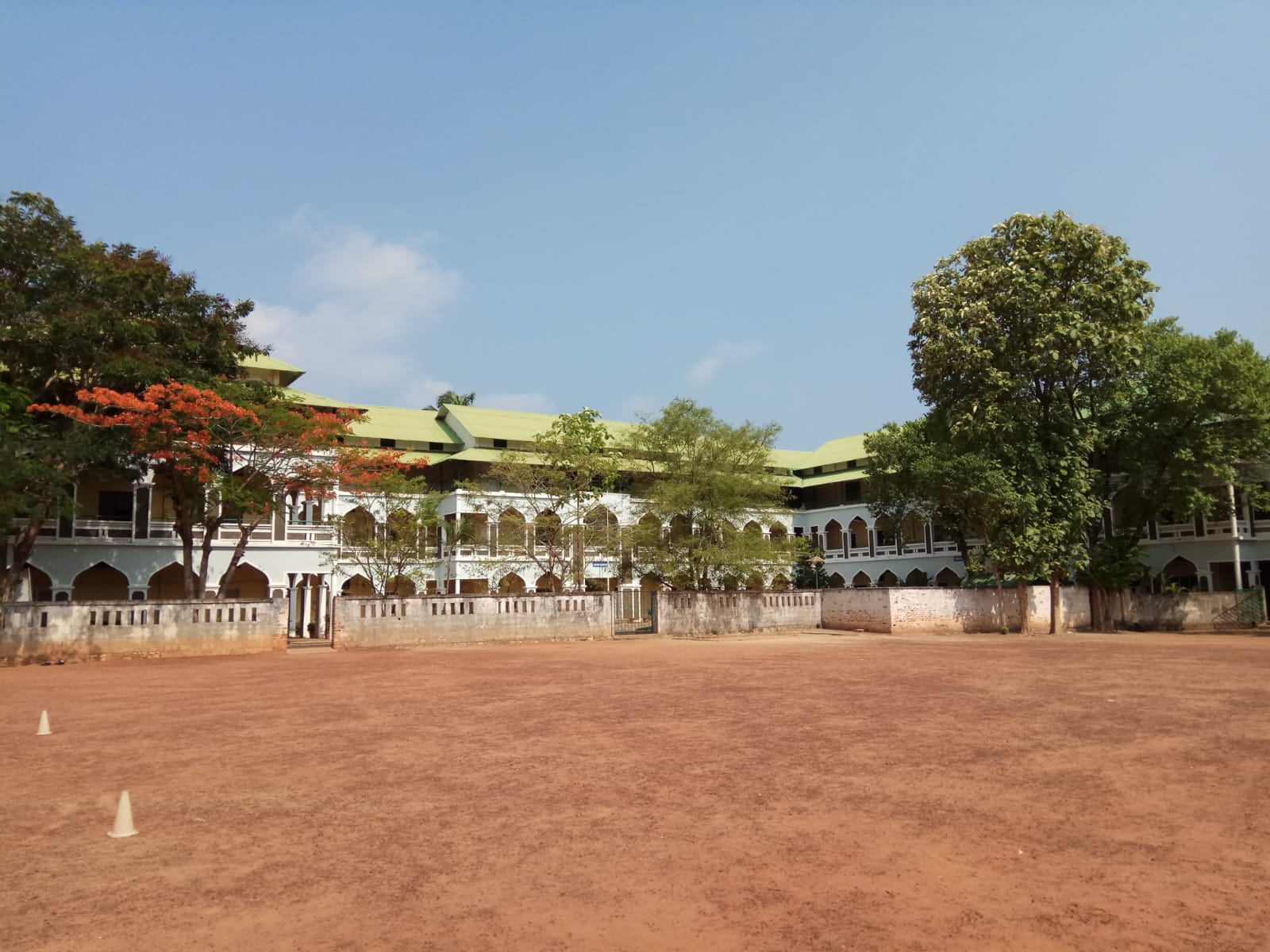 A two-story educational or institutional building with distinctive Islamic architecture featuring multiple arched doorways and windows across both levels. The structure has a light green or yellow-green roof and white walls with decorative arches. A low wall with balustrades runs along the front of the building. The foreground shows a large red dirt sports ground or playground with cone markers. Mature trees, including one with reddish-orange foliage, frame the building on both sides against a clear blue sky.