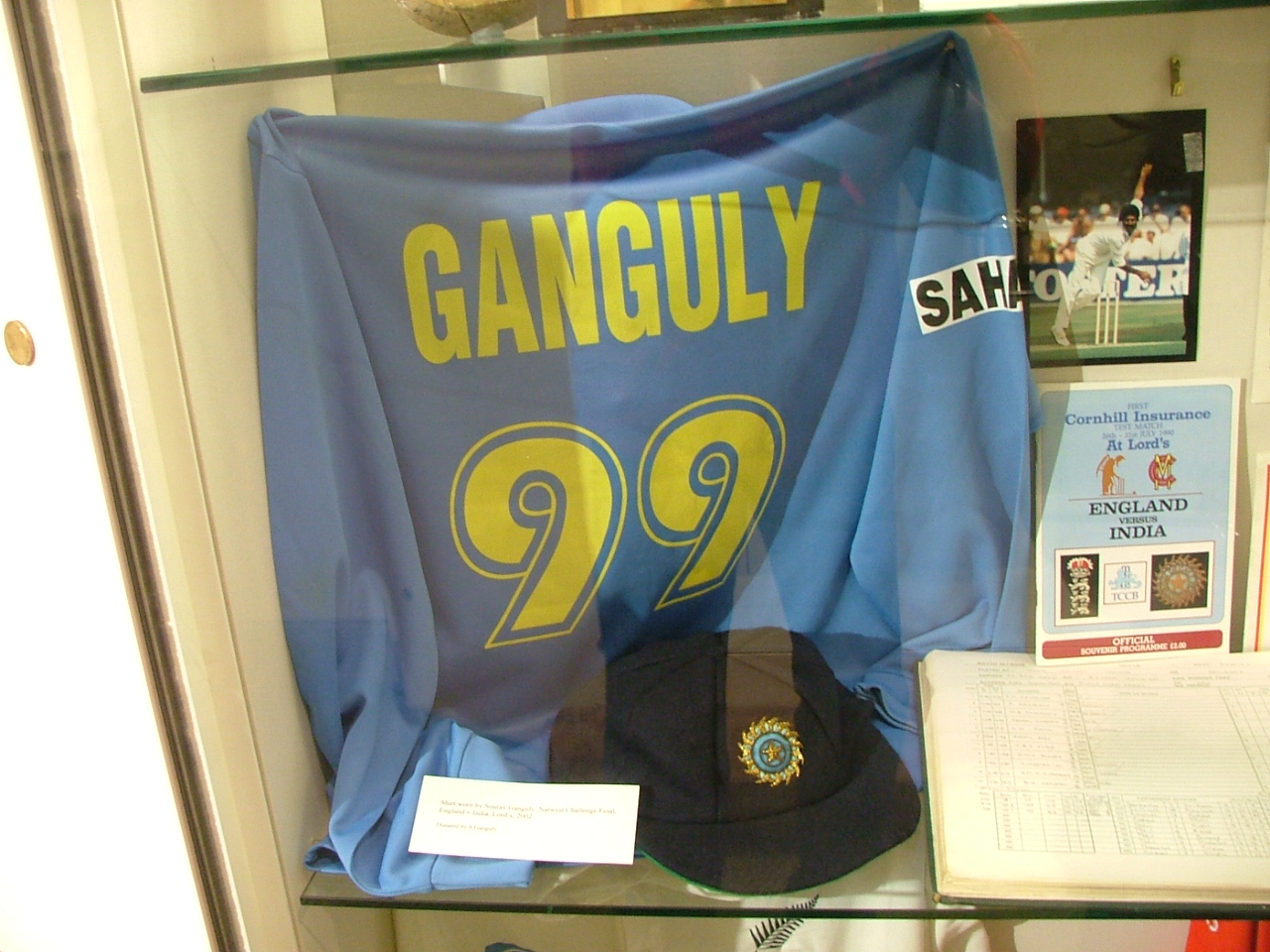 A blue coloured T-shirt displayed at a store window. The T-shirt has the words "Ganguly" and the number 99 below it, both in yellow colour. Beside the T-shirt, a picture and an open book is visible.