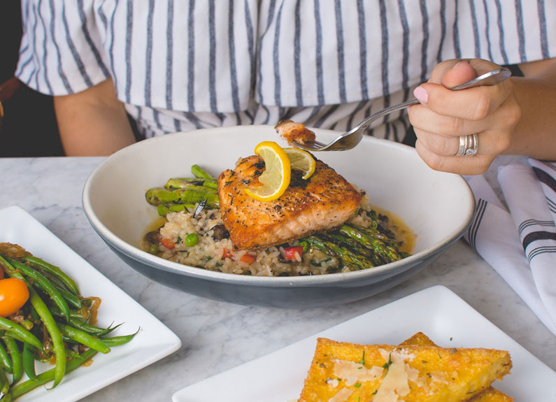 a hand holding a fork about to cut a salmon filet on a bed of rice on a white plate