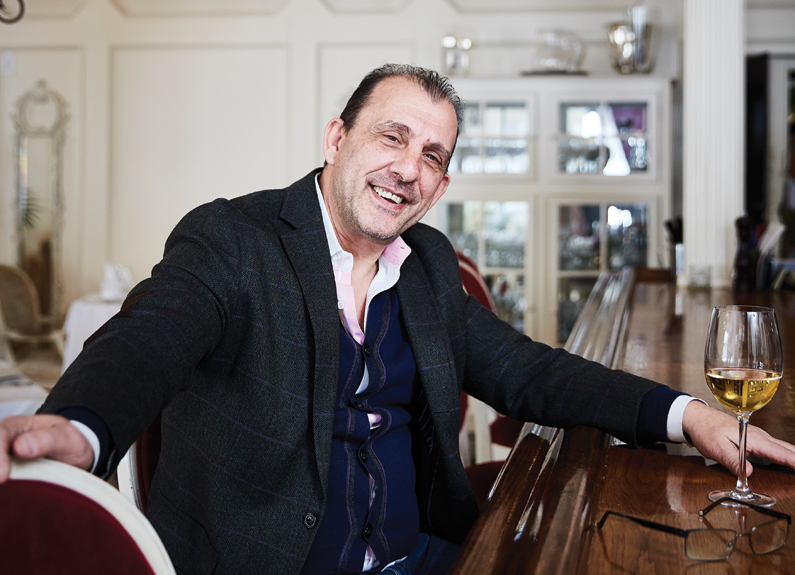 a smiling man sitting at a bar with a glass of white wine