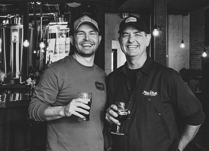 two smiling menin baseball caps holding beers and standing in front of brewing equipment