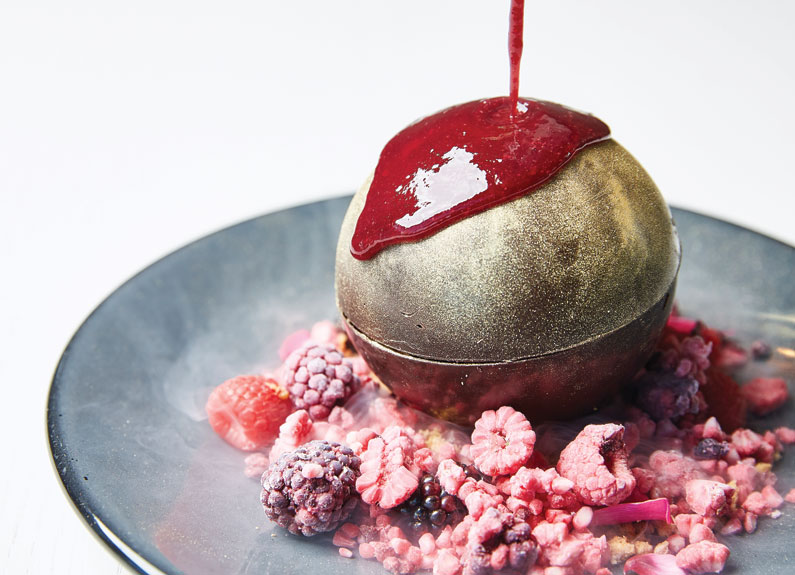 a dessert consisting of a chocolate orb atop freeze-dried berries being drizzled with berry sauce