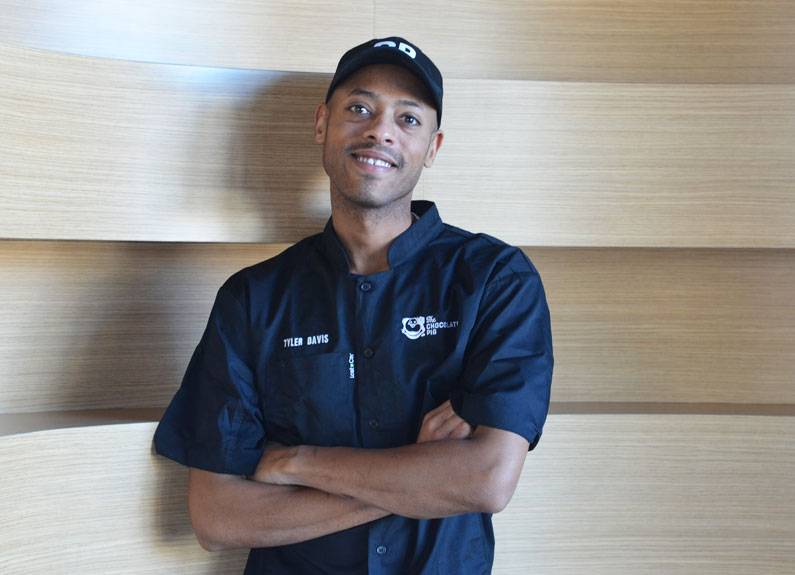 a man in a blue chef coat leaning against a wood wall