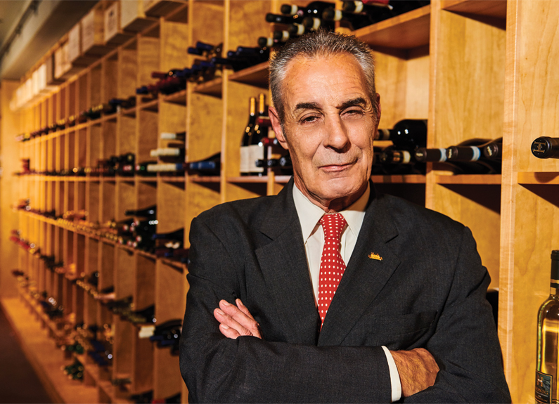 a man in a suit crossed arms standing in front of a wall of wine