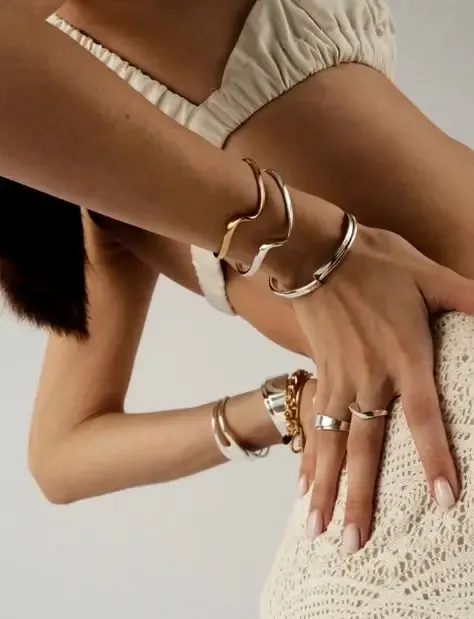 A woman in a beige crochet skirt and top poses with her arm bent, displaying elegant gold and silver bracelets and rings on her hand. The mood is stylish and serene.