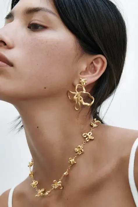 Close-up of a woman wearing a striking gold necklace and matching abstract earrings. Her expression is serene, and she has dark hair tied back.