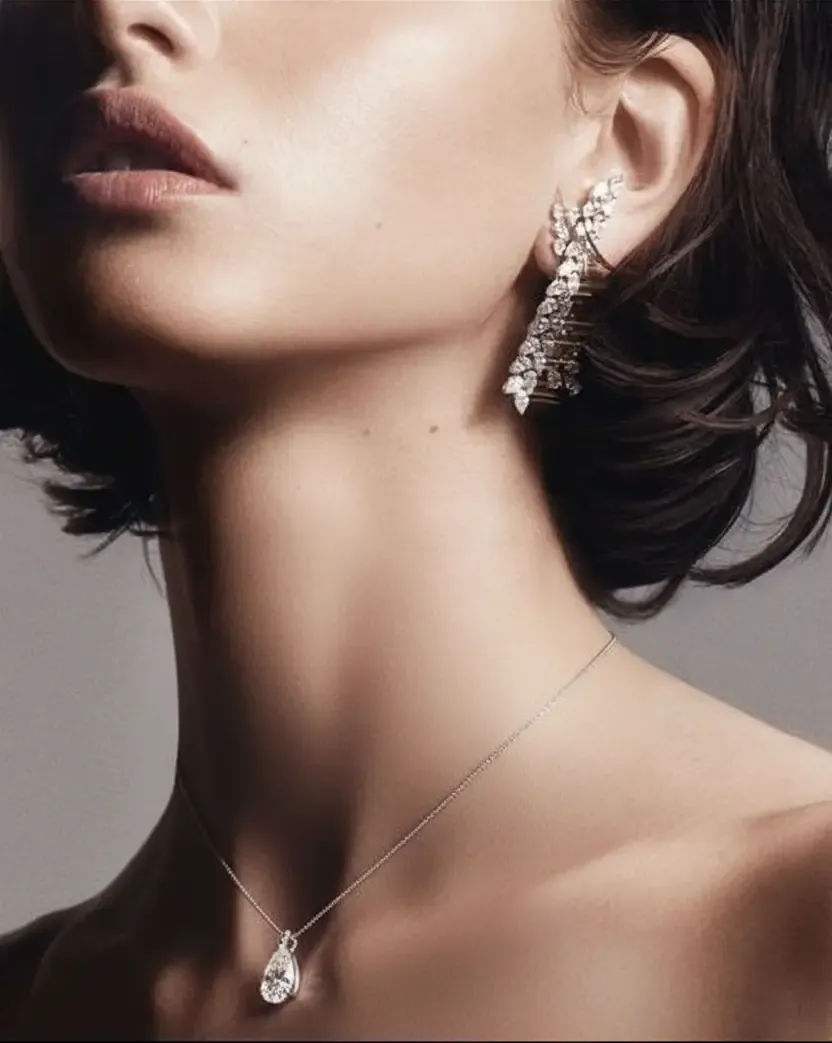 Close-up of a woman wearing luxurious diamond jewelry, including a large, intricate earring and a teardrop pendant necklace against smooth skin.