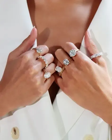 Hands adorned with multiple gold rings featuring large diamonds, elegantly displayed against a white blazer. The image conveys luxury and sophistication.