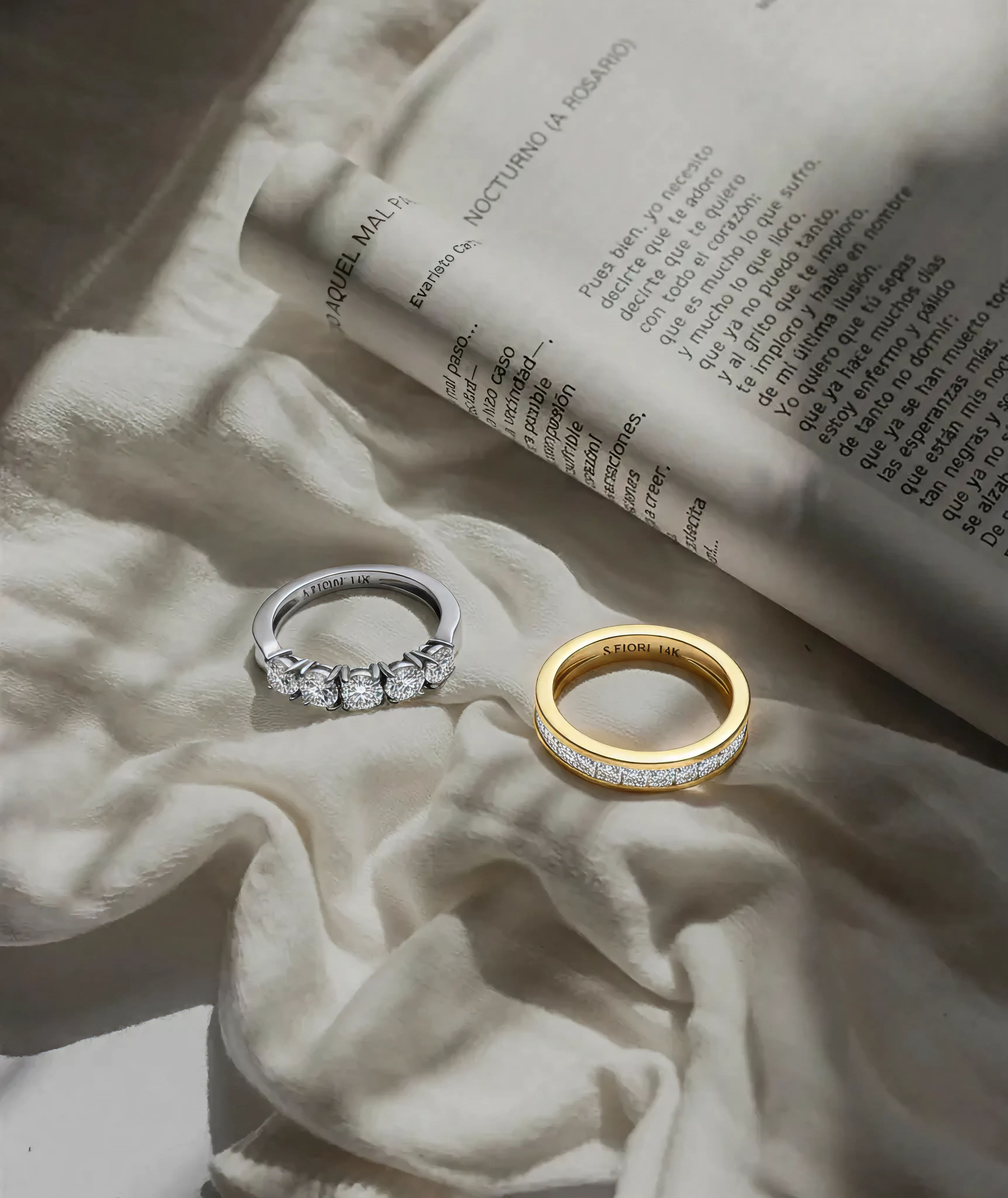 Two rings on soft fabric: a silver ring with multiple diamonds and a gold ring with embedded gems. An open book casts a gentle shadow.