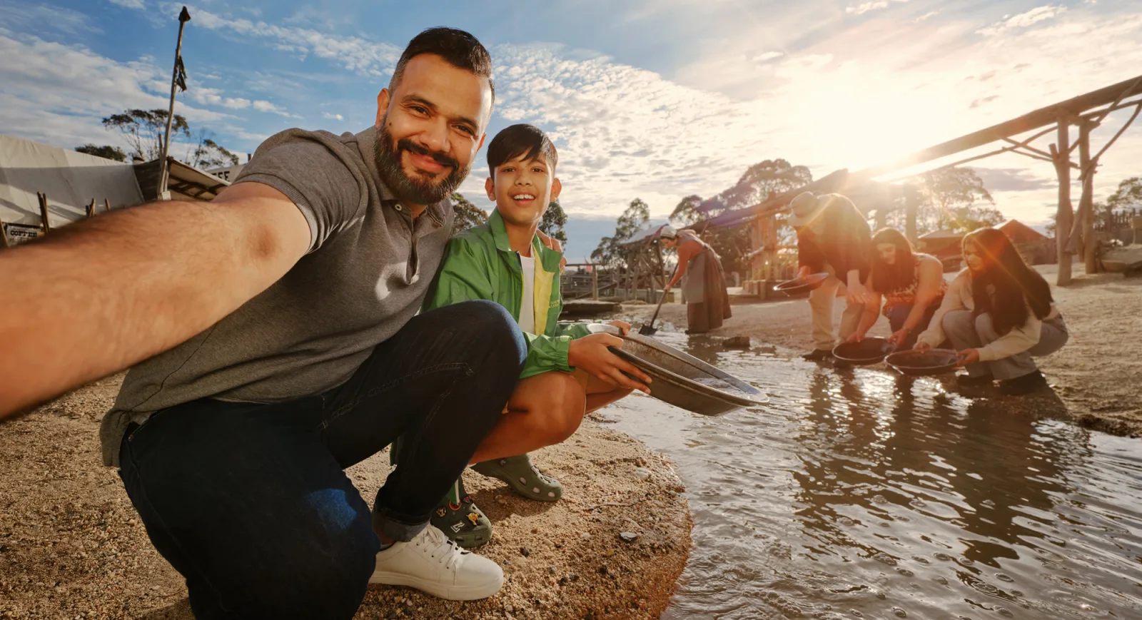 At Sovereign Hill, a man and boy capture a joyful selfie while gold panning along a sunlit creek, surrounded by other visitors engaged in the hands-on activity. Historic gold rush-era buildings line the background under a bright blue sky, highlighting the interactive, family-friendly charm of Sovereign Hill’s living history experience.