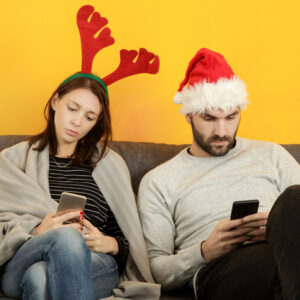 Couple wearing a santa hat and reindeer antlers sit on the couch staring at their phones