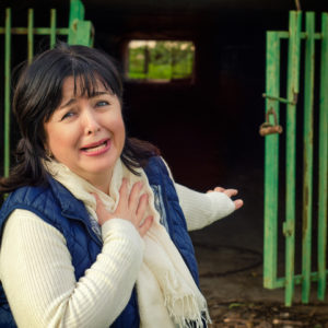 A middle-aged woman looking afraid with hand over heart stands in front of the open gates to a tunnel