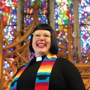 Rev Jes Kast with stained glass windows behind and a rainbow stole over shoulders