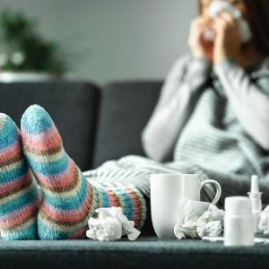 Sick woman with flu, cold, fever and cough sitting on couch at home wearing wool sock surrounding by tissue