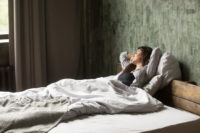 Smiling female putting her hands behind head lying and resting with closed eyes in bed.