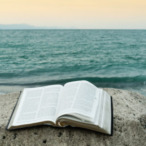 Open Bible on top of a rock in front of the sea of greenish waters and an orange sky.