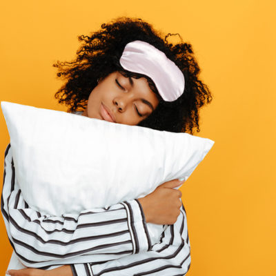 Young woman in pajamas is hugging a pillow, on a yellow background.