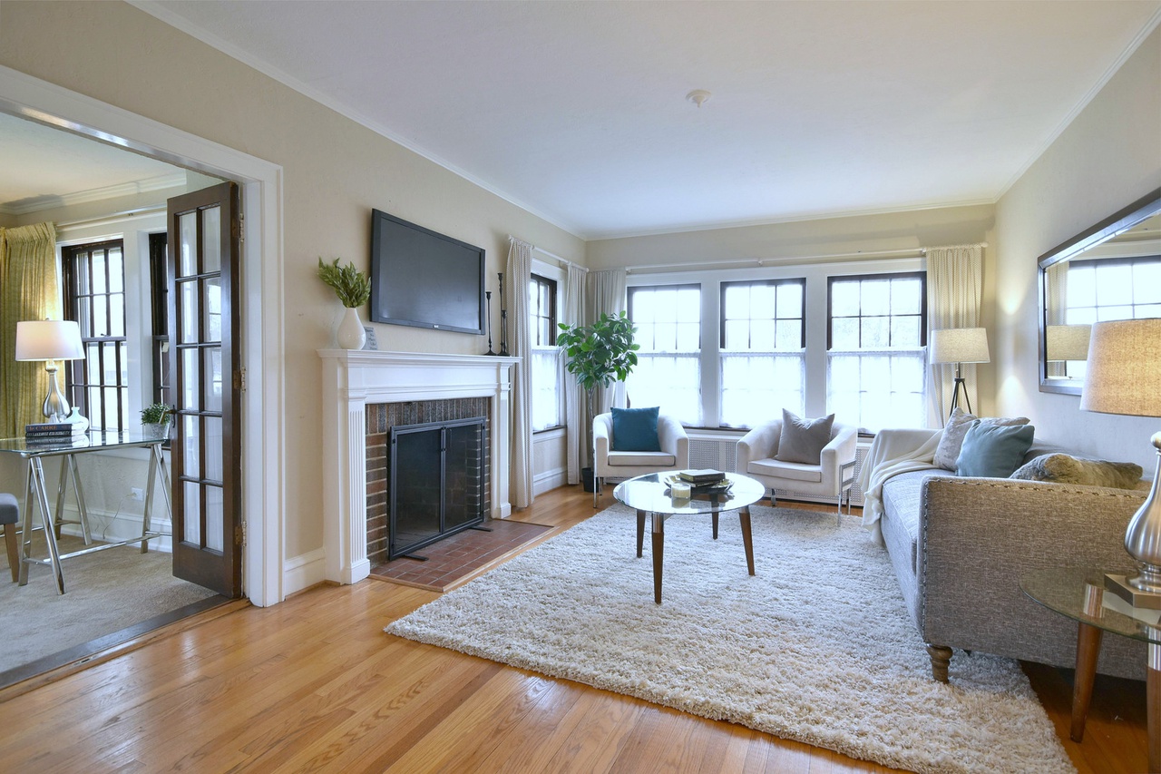 After photo of the Elmhurst single family home's living room and sunroom, beautifully staged to highlight the transitional style, featuring a faux TV, a white area rug, a glass coffee table, and comfortable seating arranged to invite conversation. The sunroom now includes a functional workspace with tasteful accessories.