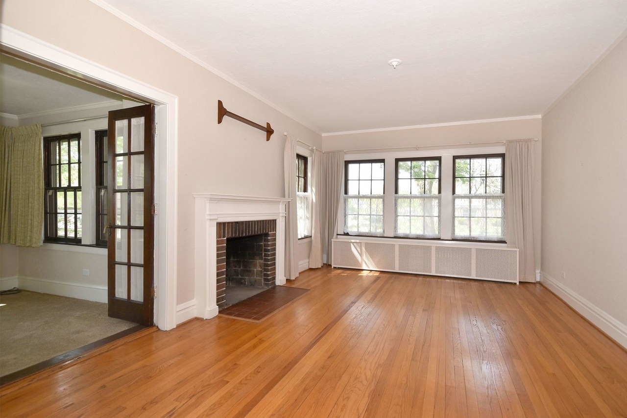 Before photo of the Elmhurst single family home's living room and sunroom, empty and awaiting transformation, showing hardwood floors, cream walls, a fireplace, and the potential for a cozy, inviting space.