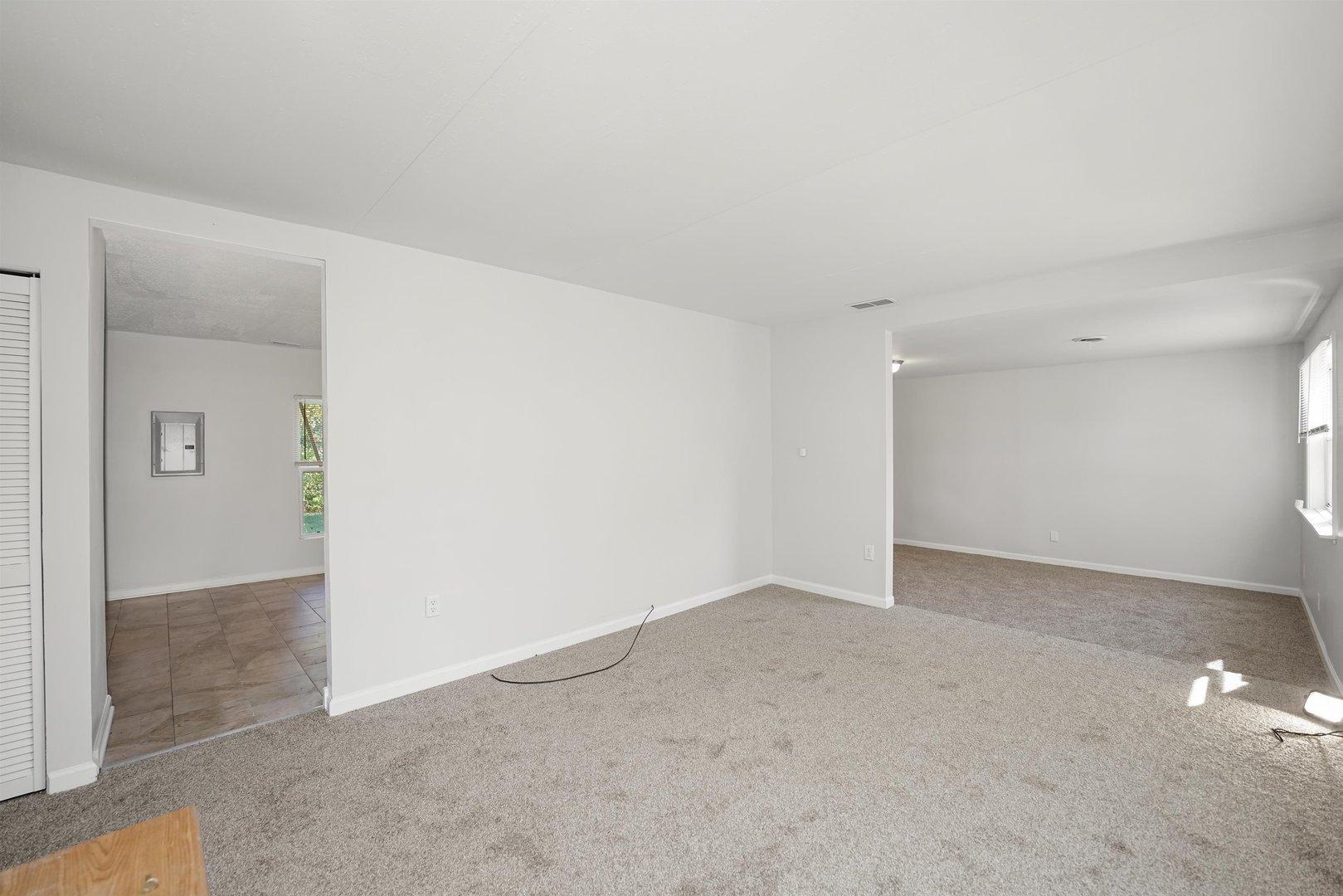 Before staging, the living room, kitchen, and dining room in Richton Park property, showing the space's initial condition with white painted walls, carpeted living and dining room, and tiled kitchen.