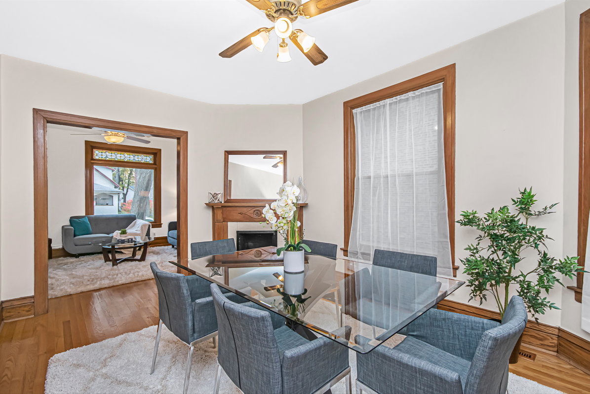 After being staged, this same Evanston home's dining room and living room have transformed into elegant, comfortable spaces. The dining room now has a large glass dining table with contemporary chairs, a white area rug, a faux tree, an orchid centerpiece, and new window treatments. The living room has mid-century-style love seat and sofa, a white area rug, a wood coffee table with a round glass top, decorative accessories, lamps, and throws on the sofa/love seat. This photo is part of a before/after series showcasing the power of home staging.