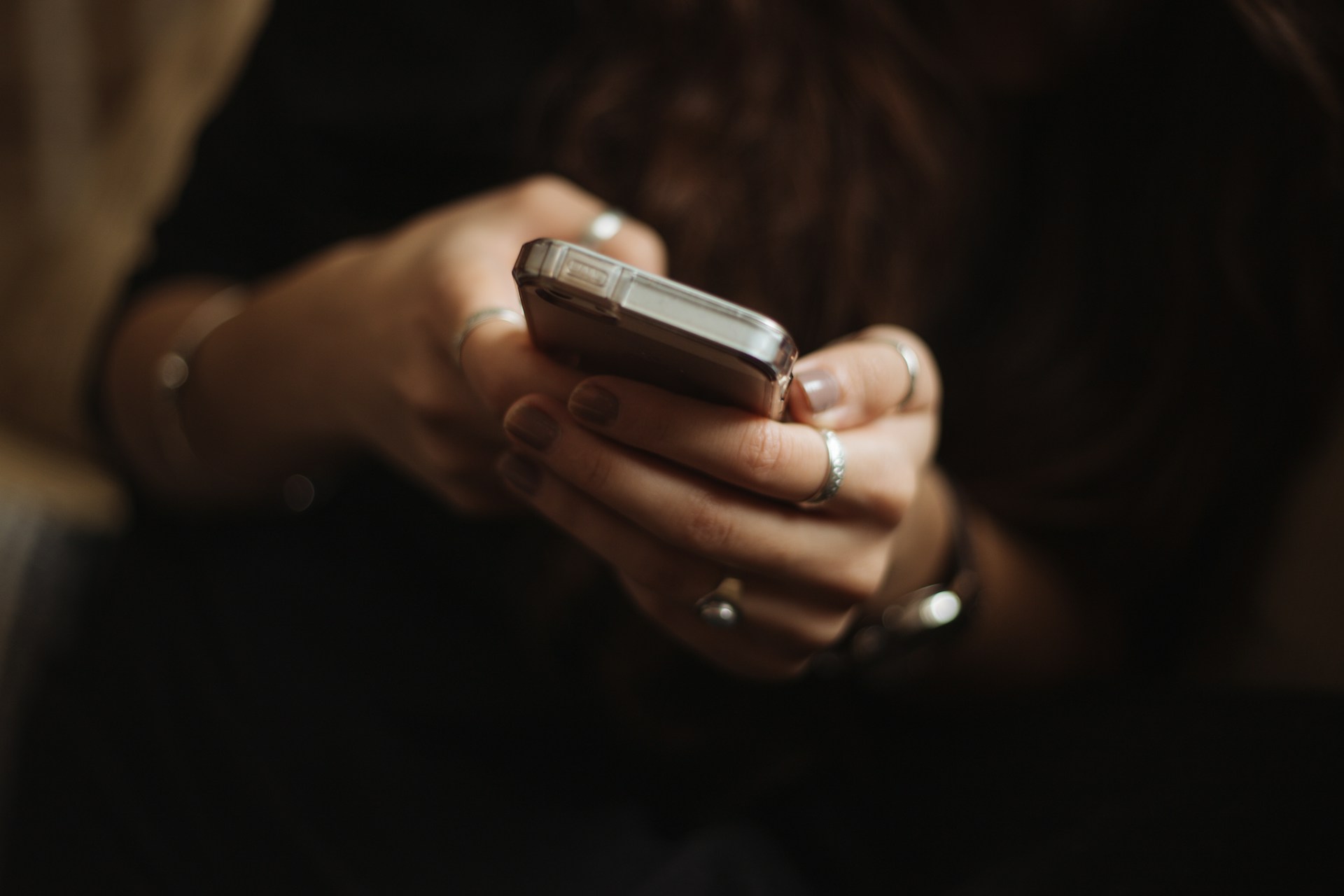 Close-up of a person's hands holding a smartphone in a dark, low-light setting, rings visible on several fingers.
